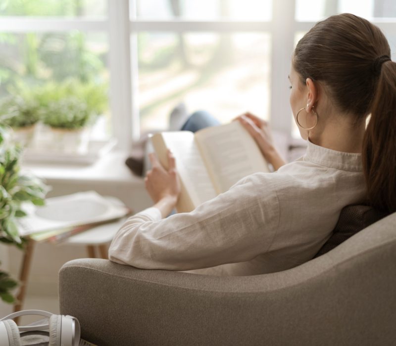 Young woman relaxing at home next to a window, she is sitting on the armchair and reading a book